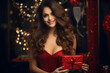 © manof - soft glow of twinkling lights in a dimly lit room, a woman holds a crimson gift box adorned with a festive ribbon.