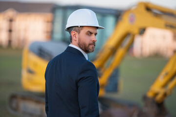 Wall Mural - Construction owner near excavator. Confident construction owner in front of house. Architect, civil engineer. Man construction owner with a safety vest and hardhat at construction site.