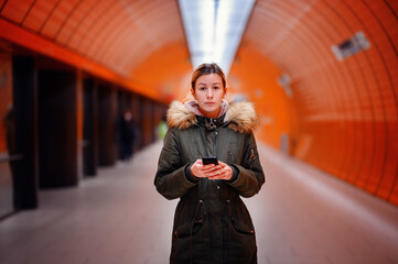 Wall Mural - Woman Texting in Orange Subway Corridor, Focused young woman texting on phone in a vibrant orange-lit subway tunnel