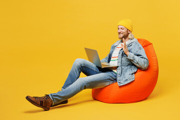 Wall Mural - Full body fun young IT man he wears denim shirt hoody beanie hat casual clothes sit in bag chair hold use work on laptop pc computer waving hand isolated on plain yellow background. Lifestyle concept.