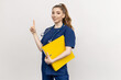 © makedonski2015 - A healthcare worker in blue scrubs holding important documents while standing against a blank white wall.The nurse points her finger to the side. Mockup