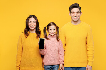 Poster - Young smiling parents mom dad with child kid girl 7-8 years old wear pink sweater casual clothes hold use blank screen area mobile cell phone isolated on plain yellow background. Family day concept.