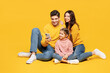 © ViDi Studio - Full body young smiling happy parents mom dad with child kid girl 7-8 years old wear pink sweater casual clothes sitting use mobile cell phone isolated on plain yellow background. Family day concept.