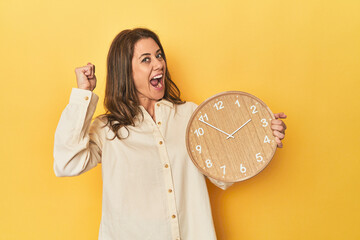 Wall Mural - Woman holding clock on yellow backdrop raising fist after a victory, winner concept.