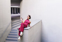 Smiling Woman Wearing Pink Jumpsuit And Sitting On Wall Near Staircase