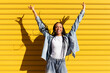 © VISTA by Westend61 - Cheerful woman standing with arms raised in front of yellow shutter