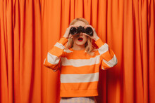 Woman Looking Through Binoculars Standing In Front Of Orange Curtain