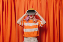 Surprised Woman Wearing Virtual Reality Simulator Standing In Front Of Orange Curtain