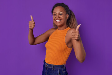 Wall Mural - Young laughing beautiful African American woman in casual clothes shows thumbs up and looks at camera recommending you to purchase advertised product posing on isolated purple background.