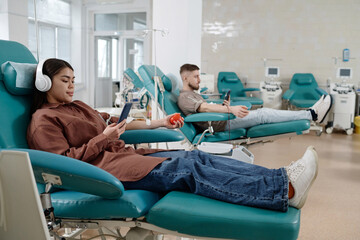 Wall Mural - Blood donors sitting side by side on medical recliner chairs in hospital and browsing smartphones