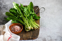 Fresh Arugula Leaves Neatly Bundled On A Wooden Cutting Board Next To A Bowl Of Pink Himalayan Salt And A Cloth Napkin, Ready For Salad Preparation On A Textured Kitchen Table