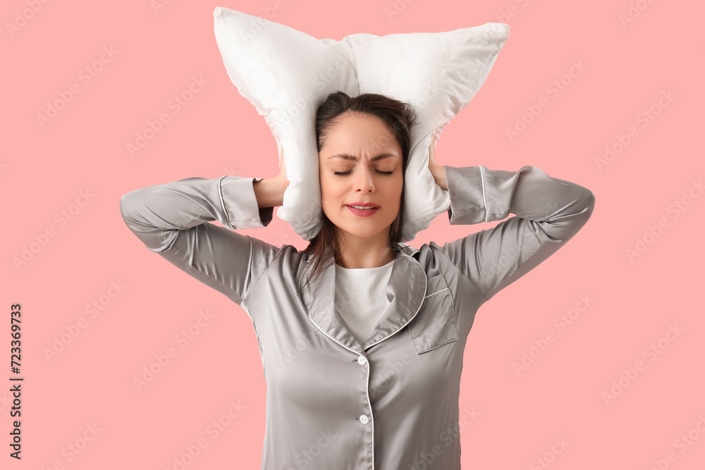 Displeased young woman with pillow plugging ears on pink background