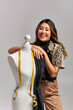 © LIGHTFIELD STUDIOS - cheerful asian stylist looking at camera near mannequin with fabric and measuring tape on grey