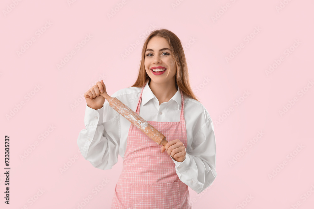 Pretty young woman in apron with rolling pin on pink background