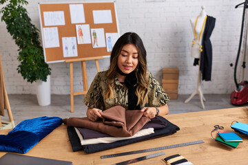 pleased asian personal stylist checking quality of fabrics samples on work desk in own atelier