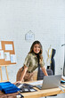 © LIGHTFIELD STUDIOS - joyful asian personal stylist looking at camera at work desk with laptop in atelier, small business