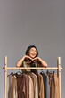 © LIGHTFIELD STUDIOS - excited asian clothes designer laughing with closed eyes near rack with trendy clothes on grey