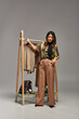 © LIGHTFIELD STUDIOS - fashionable asian clothes designer standing near rack with trendy bespoke clothes on grey backdrop