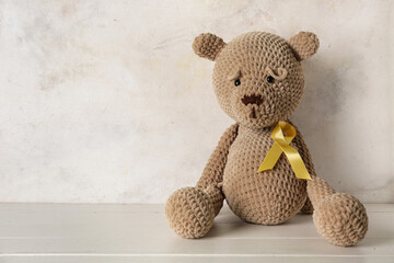 Wall Mural - Toy bear with golden ribbon on white wooden table against grunge background. Childhood cancer awareness concept