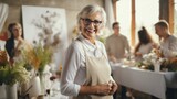 Fototapeta  - An enthusiastic elderly woman with glasses smiling joyfully, hosting a flower arrangement workshop in a bright setting.