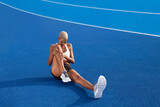 Fototapeta Koty - An African American female athlete engages in warm-up exercises, sitting on the Olympic blue track, embodying the concept of race training and dedication in sports