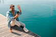 A trendy guy is sitting on a dock near sea and waving at the phone during his video call on the phone.
