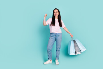 Full length photo of cute lucky mixed race woman wear pink t-shirt rising fist holding bargains empty space isolated turquoise color background