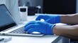 A person wearing blue gloves is typing on a laptop. This image can be used to illustrate concepts such as technology, work, cybersecurity, or data entry