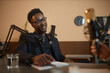 Medium shot of friendly Black man in eyeglasses and suit listening to unrecognizable podcast host question while sitting in studio