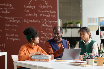 Wall Mural - Front view portrait of senior Black teacher talking to students at table in college classroom and pointing to laptop screen copy space