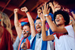 © Drazen - Happy black woman cheering with group of fans during sports match at stadium.