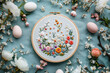 Flat lay postcard with a hoop with beautiful flowers embroidery, on a table, surrounded by colorful Easter eggs painted in different colors to celebrate Easter. Top down view.