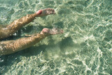 Fototapeta Łazienka - Woman swimming on the sea. Female legs under clear sea water. Realistic top view photo of a women's legs barefoot. Relax woman lying in turquoise crystalline blue water on beach vacation summer.