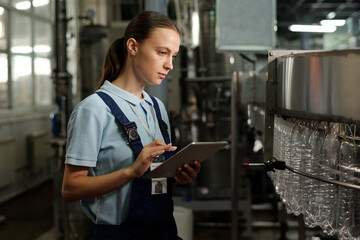Poster - young quality control staff with tablet looking at empty plastic bottles in factory conveyor and ent