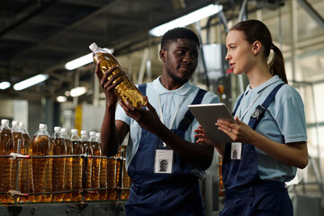 Leinwandbilder - Young female inspector with tablet talking to male colleague holding packed plastic bottle with carbonated lemonade or soda