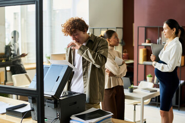 Poster - Yawning office manager standing by copier machine and looking through financial paper among intercultural colleagues in coworking space