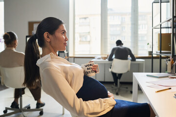 Poster - Young bored pregnant businesswoman in smart casualwear having tea or coffee while sitting in front of computer screen in office