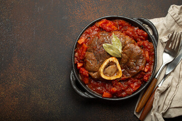 Wall Mural - Traditional Italian dish Ossobuco all Milanese made with cut veal shank meat with vegetable tomato sauce served in black casserole pan top view on rustic brown background, copy space.