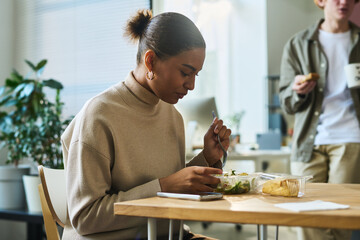 Poster - Young hungry African American female white collar worker sitting by table at lunch break and having vegetarian salad from plastic container