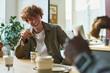 © pressmaster - Young laughing male office worker with cookie or some other snack in hand sitting in front of colleague and enjoying lunch with him