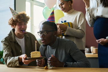 Poster - Young businessman blowing burning candle on piece of tasty birthday cake after making wish while sitting among happy colleagues