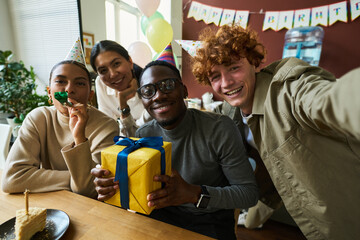 Poster - Young intercultural white collar workers with noisemakers and gift looking at camera while cheerful businessman taking birthday selfie