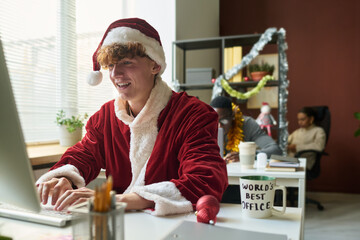 Leinwandbilder - Happy young businessman in costume of Santa Claus typing on keyboard of desktop computer while sitting by workplace against colleagues