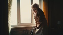 Sad Elderly Woman Standing With A Walking Frame Near The Window, Feeling Lonely