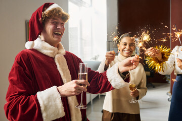 Poster - Young laughing businessman in costume of Santa Claus holding burning sparkler and flute of champagne while enjoying xmas celebration