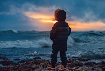 Poster - Boy in warm winter clothes standing on seashore during storm in winter at sunset