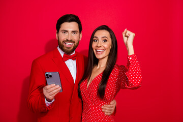 Poster - Photo of two attractive delighted lovers embrace hold smart phone raise fist empty space isolated on red color background