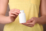 Fototapeta  - Woman holding blank white bottle with vitamin pills against light brown background, closeup