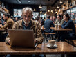 © Fabio Levy - Old Man sitting at a table with a laptop and a cup of coffee. She is focused on her work. There are other people in the background, engaged in their own activities.