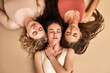 Feminine sensuality. Top view of females trio with dark curly, blond short and straight long hair lying head to head on beige background. Charming women wearing comfy sport bra expressing self love.